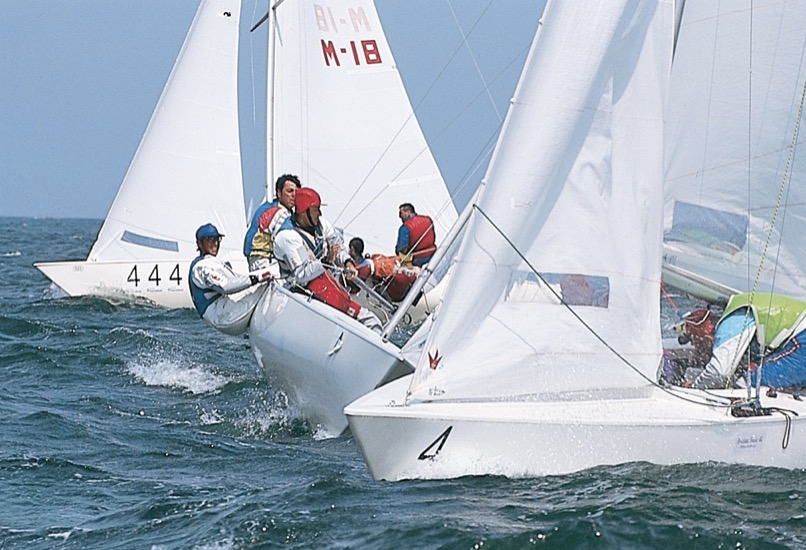 Das Boot im Vordergrund wendet gerade. Gegen den Wind laufend sind die Segel drucklos. Mit den Segeln wechselt auch die Crew die Seite. Kurz zuvor lief das Boot auf Backbordbug (wie das Boot dahinter). Nach der Wende läuft es auf Steuerbordbug (wie das Boot 444 im Hintergrund) weiter.